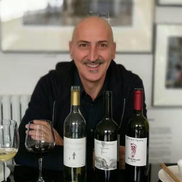 Simone Pezzatini sorride seduto a un tavolo con tre bottiglie di vino e due calici, simbolo della sua esperienza nel settore vitivinicolo